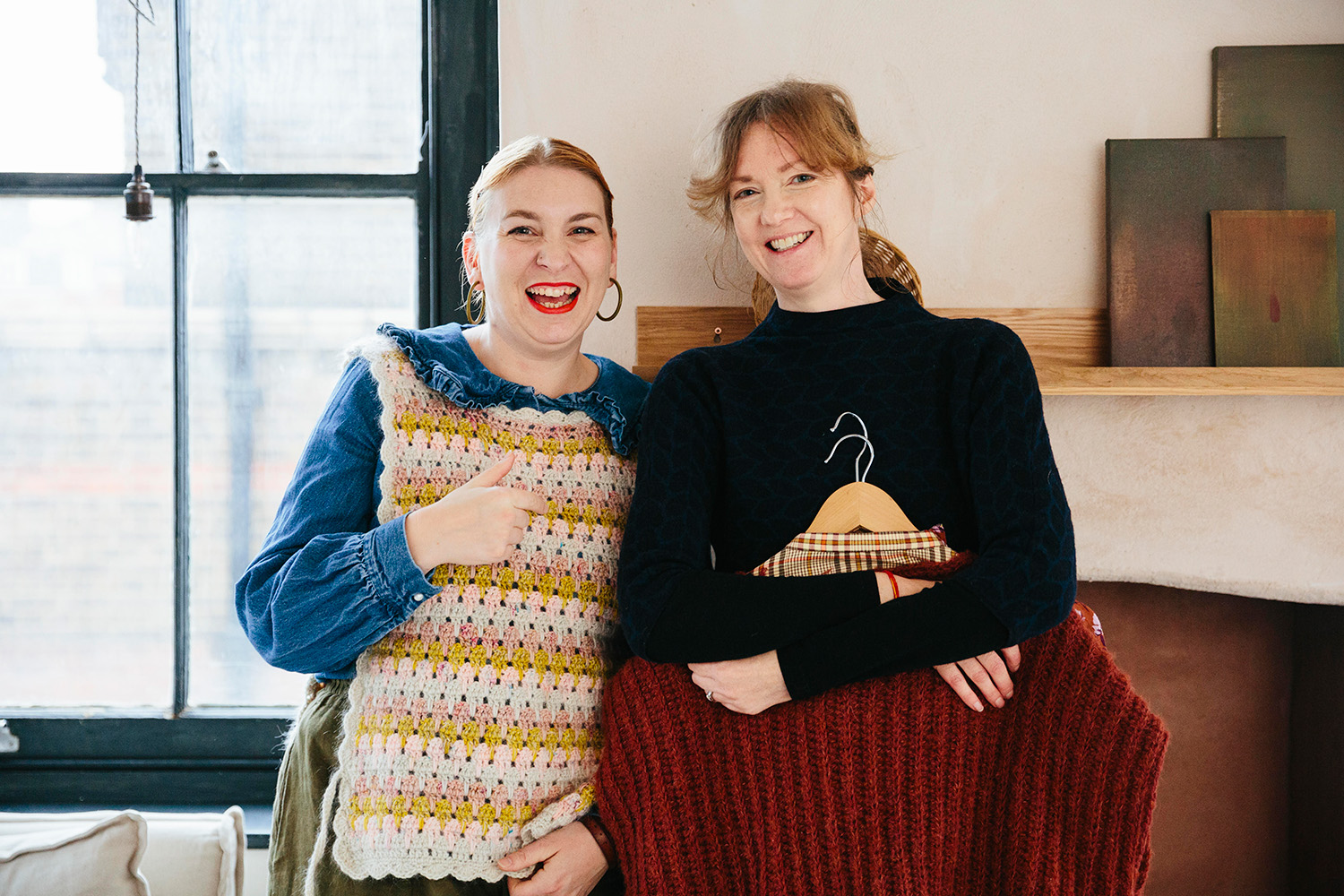 Two women showcasing handmade crochet and knitwear indoors