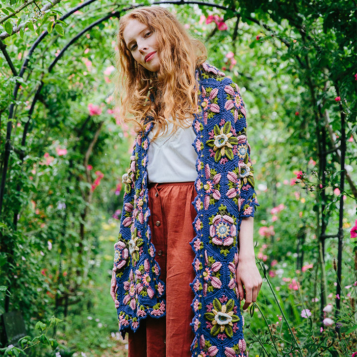 Model standing in a garden wearing a detailed crochet shawl with floral motifs – Climbing Rose design.