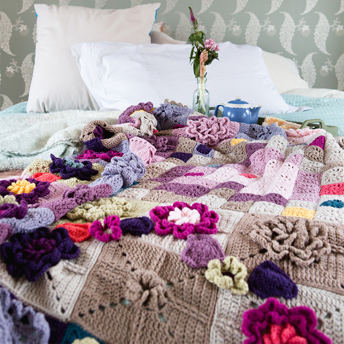 Intricate crochet blanket with 3D floral motifs in soft pinks and purples on a bed – Mystery Blanket by Sabina Poonwassie.