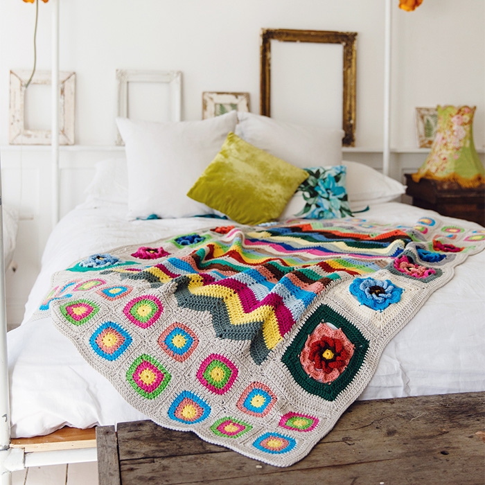 Bright and playful crochet blanket with zigzag and floral motifs draped over a white bed – design by Annelies Baes.